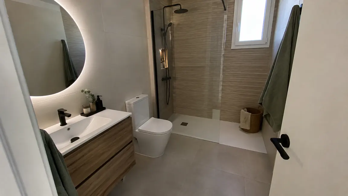 Baño reformado por Reformas DLCP con ducha de obra, mampara de vidrio, mueble suspendido de madera y espejo redondo retroiluminado