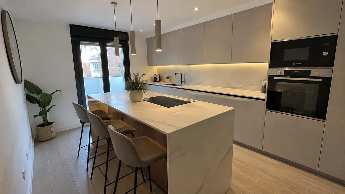 Cocina reformada por Reformas DLCP con isla, encimera porcelánica veteada, muebles claros sin tirador y columna de horno y microondas