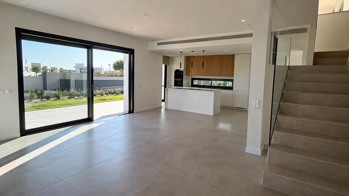 Salón y cocina de una vivienda de obra nueva terminada por Reformas DLCP, con isla, ventanal corredero al jardín y escalera con barandilla de vidrio
