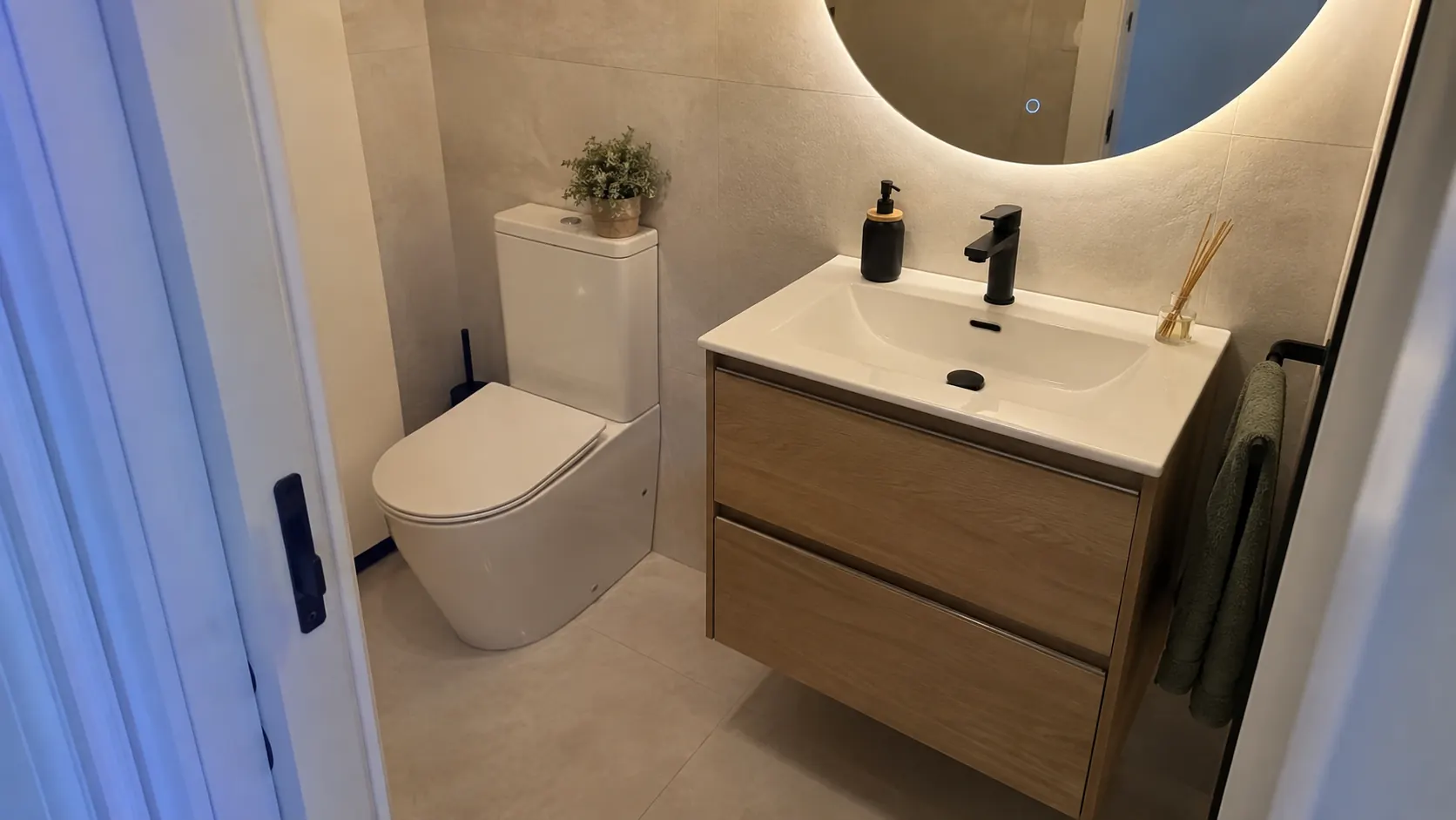 Aseo reformado por Reformas DLCP con inodoro, mueble de lavabo suspendido en madera y espejo redondo retroiluminado